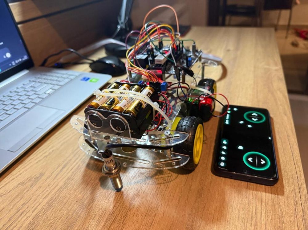 The reconnaissance robot: clear acrylic chassis, battery pack and downward-facing inductive sensor at the front, phone with the control app beside it